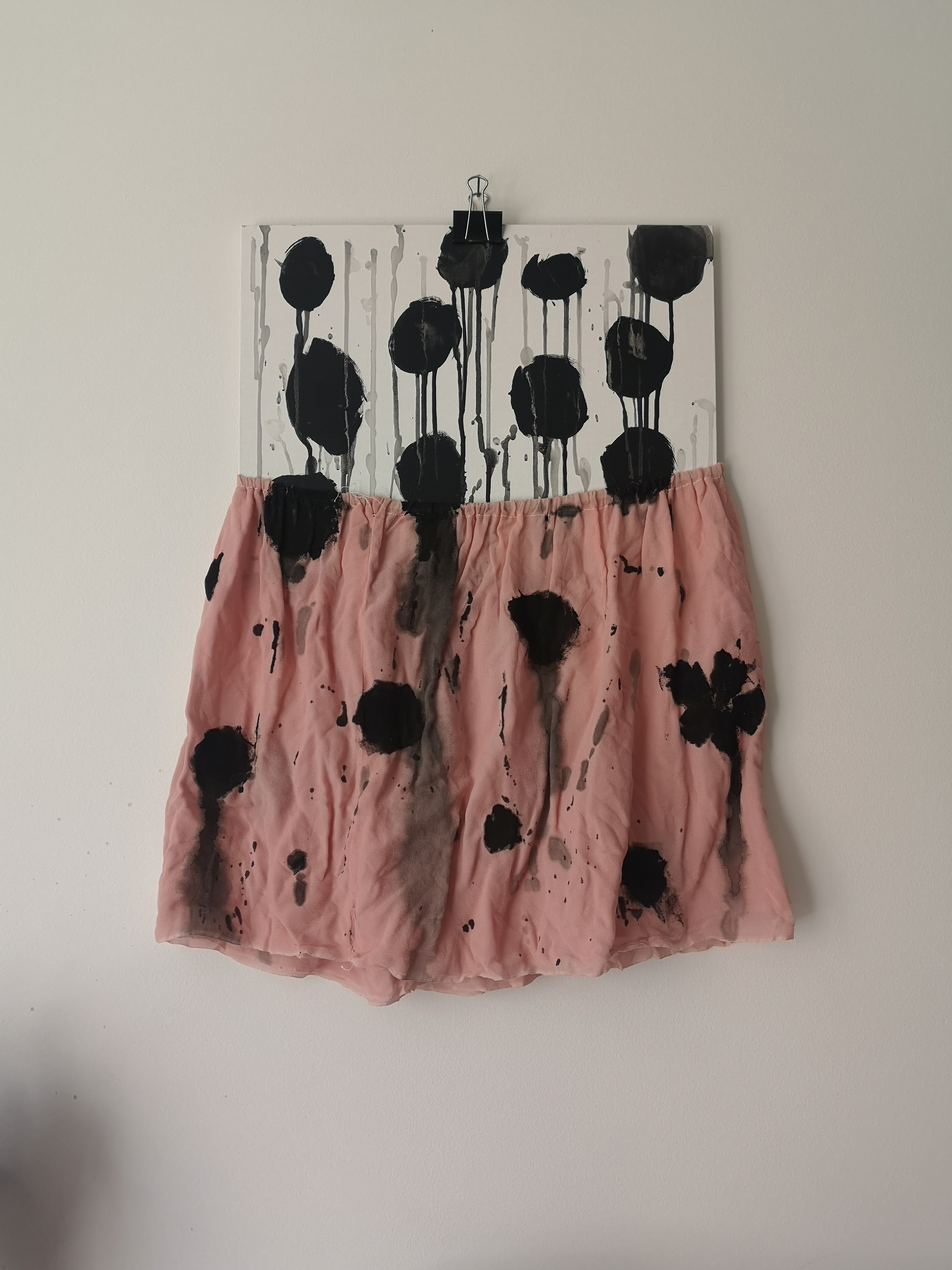 Kunstwerk aus weißer Malpappe mit einem rosa Rock an der Malpappe fixiert. Auf beidem sind große schwarze Tupfen, deren Tropfen über das Bild laufen.