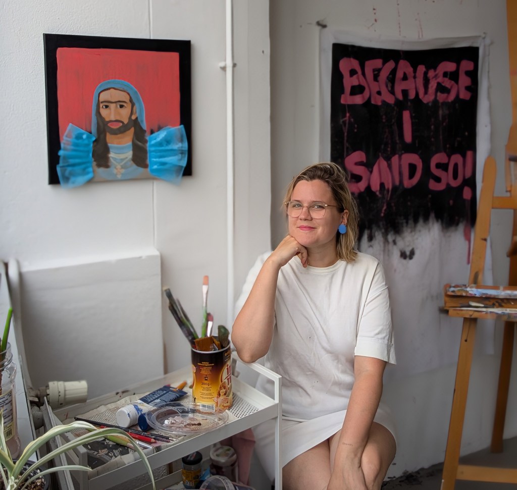 Laura Müller sitzt neben ihrem Servierwagen voller Kunstmaterialien und vor ihren Kunstwerken und ihrer Staffelei in ihrem Atlier.