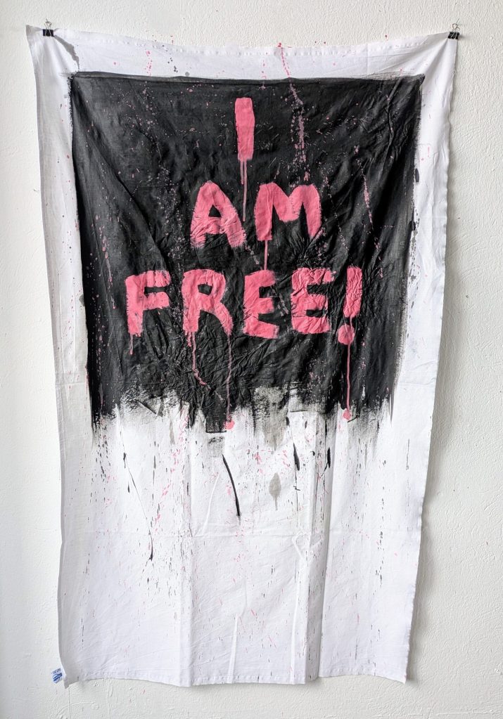 Kunstwerk, dass den Schriftzug "I am free!" in großer pinker Schrift auf schwarzem Hintergrund zeigt. beides ist mit Acryl auf DDR-Tischtuch gemalt. Das Tischtuch ist mit Farbe bespritzt und zerknittert.