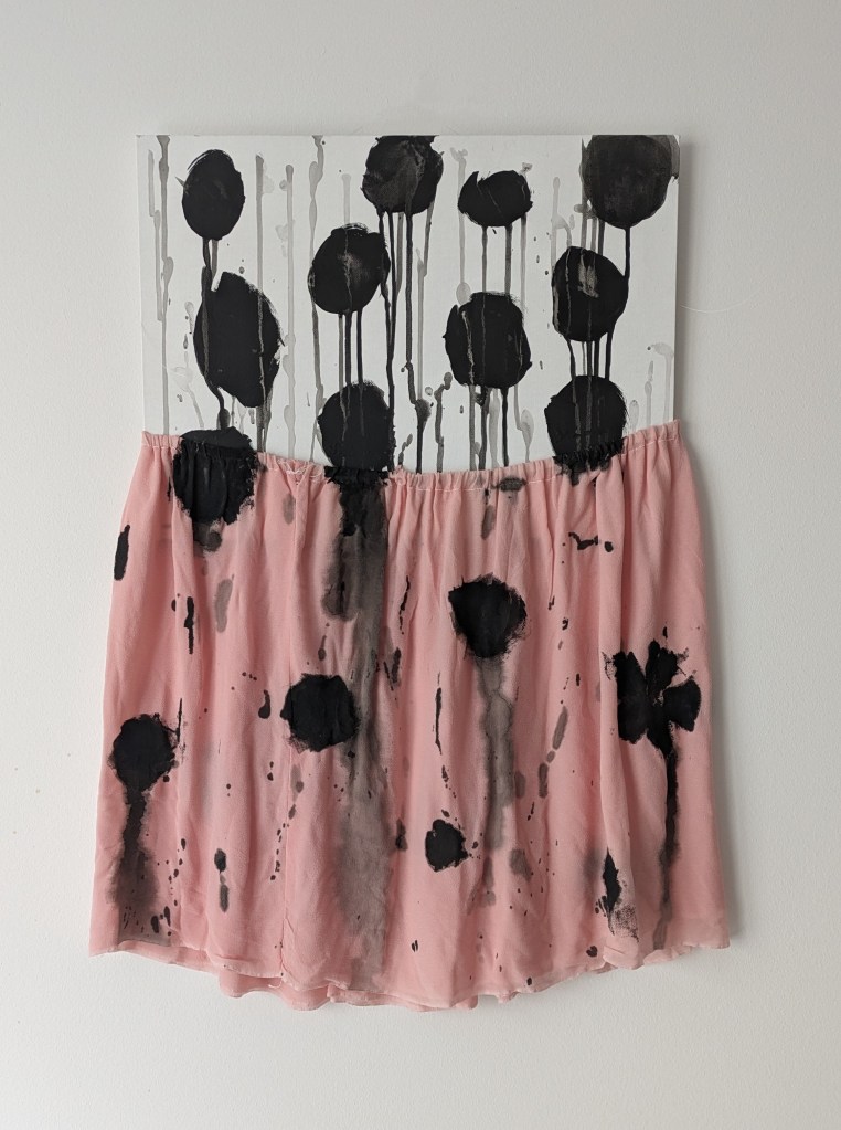 Kunstwerk aus weißer Malpappe mit einem rosa Rock an der Malpappe fixiert. Auf beidem sind große schwarze Tupfen, deren Tropfen über das Bild laufen.