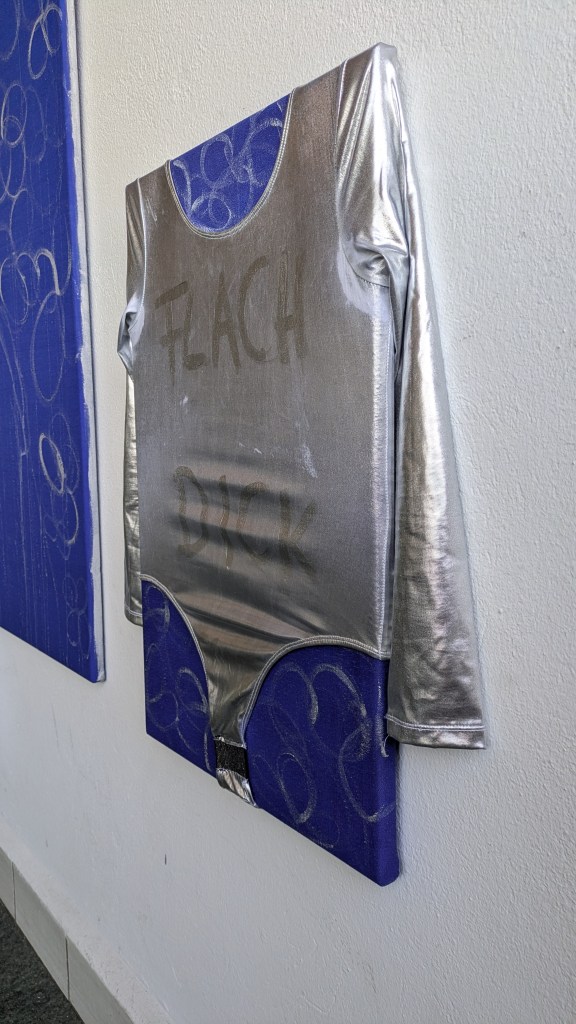 Nahaufnahme des Bildes Male Gazed. Silberner Body auf einen Keilrahmen aufgezogen. Auf ihm sind in silberner Schrift "Flach, dick, groß" zu lesen an den Stellen der Brüste, des Bauches und der Vulva.