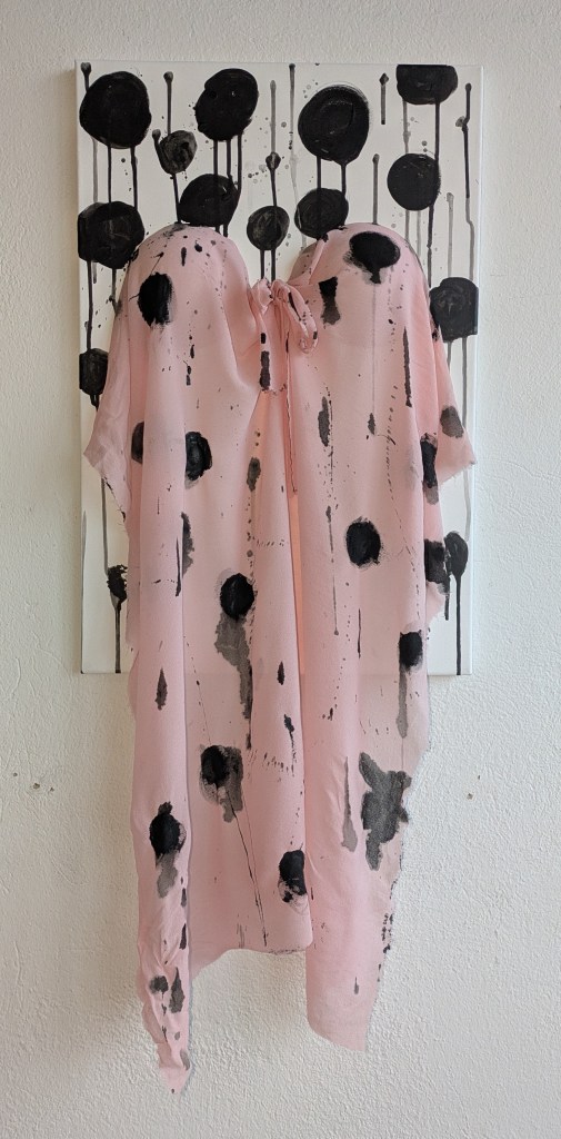 Kunstwerk aus weißer Leinwand mit einem rosa Stoff, der als Negligé anmutet an der Leinwand fixiert. Auf beidem sind große schwarze Tupfen, deren Tropfen über das Bild laufen.
