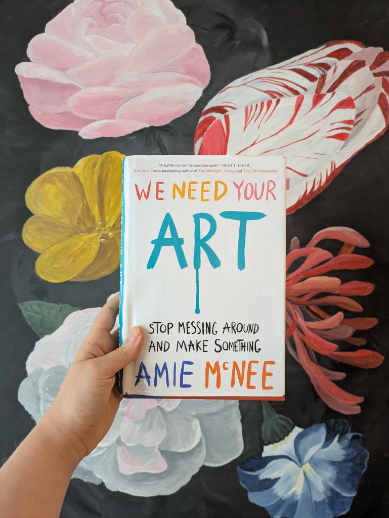 Das Buch We Need Your Art wird von einer Hand vor einem Blumenbild von Laura Müller gehaltem